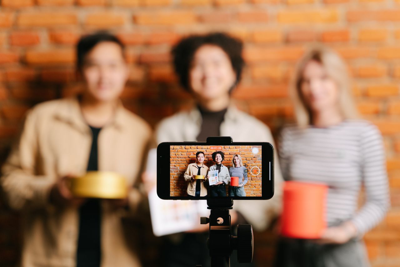 Une équipe variée de vloggers filmant une vidéo avec un smartphone devant un mur de briques en arrière-plan.